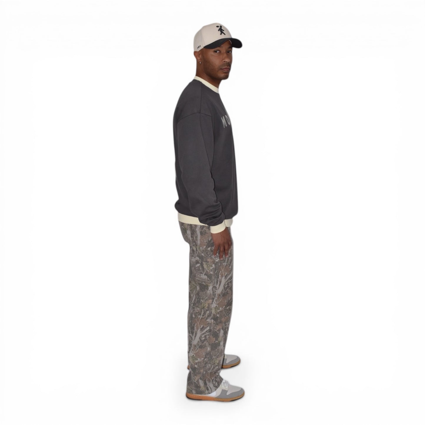 Man wearing a charcoal gray crewneck light pants, and a cap on a white background